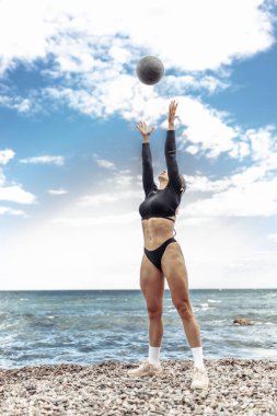 Strong athletic woman exercising with medicine ball on the beach during the day with blue sky and clouds. Functional outdoor training