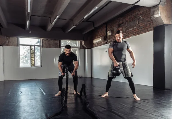 Functional training. Two male athletes are training with a kettlebell and battle ropes in a sports hall