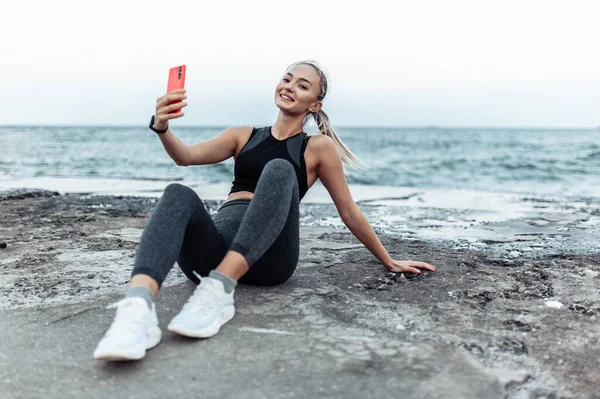 Fit woman in sportswear makes selfie on phone while sitting on urban beach
