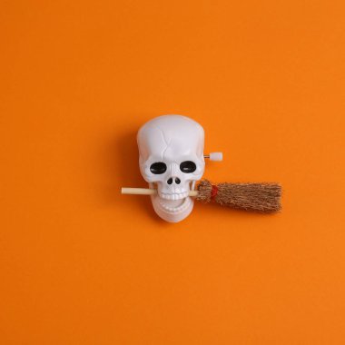 Minimal halloween still life. Skull with witch's broom on orange background. Top view
