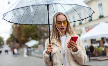 Cute fashion blonde woman in yellow sunglasses with transparent umbrella using phone in the city. Lifestyle