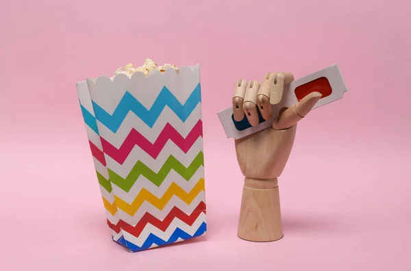 Popcorn box and wooden hand with 3d glasses on a pink background. Entertainment, going to the cinema