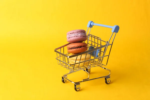 Shopping, food concept. Mini supermarket trolley with french macarons on yellow background