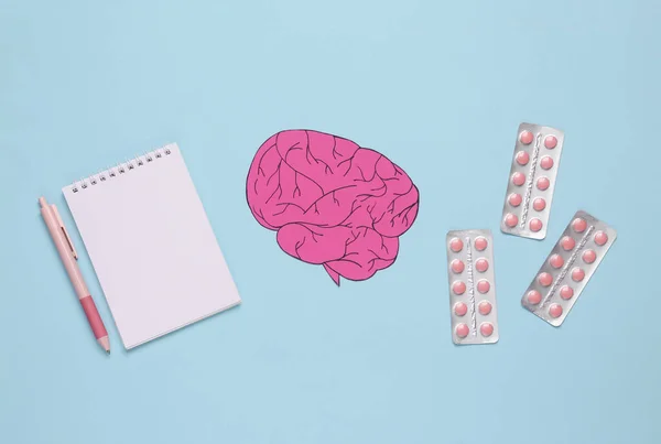 Paper-cut brain with pills and notebook on blue background. Headache treatment