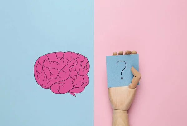Paper cut brain and memo papers with question marks in wooden hand on pink background. Finding answers to questions. Flat lay