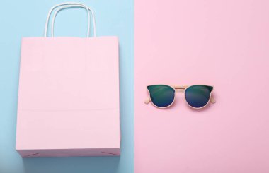 Alışveriş konsepti. Mavi-pembe pastel arka planda güneş gözlüklü kağıt alışveriş torbası. düz yat