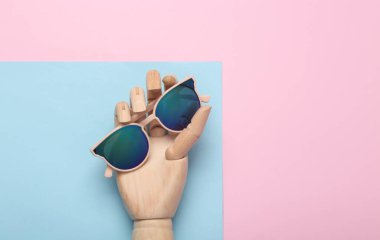 Ahşap el, mavi pastel arka planda şık güneş gözlükleri tutuyor.
