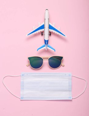 Minimum seyahat hala hayattadır. Güneş gözlüklü oyuncak uçak ve pembe arka planda tıbbi maske. Üst görünüm