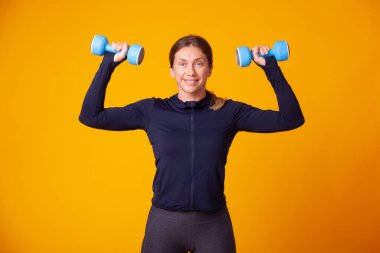 Antrenman konsepti. Spor giyinen mutlu atletik kadın, sarı arka planda elinde dambıllarla antrenman yapıyor. Vücut geliştirme ve fitness. Sağlıklı yaşam tarzı