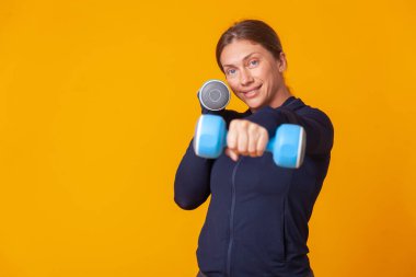 Antrenman konsepti. Spor giyinen mutlu atletik kadın, sarı arka planda elinde dambıllarla antrenman yapıyor. Vücut geliştirme ve fitness. Sağlıklı yaşam tarzı