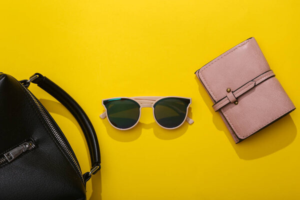 Стильные модные женские аксессуары на ярко-желтом фоне с тенью. Бумажник, сумка и солнечные очки. Плоский лежал. Вид сверху