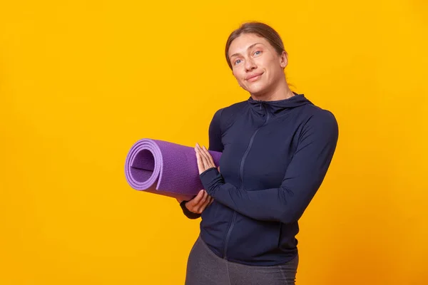 Athletic woman trainer in sportswear with a mat in her hands on a yellow studio background. Healthy lifestyle. Sport, bodybuilding and fitness concept