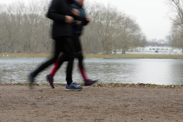 Parktaki çakıl yolunda koşan iki kişi var. Arka planda göl var.