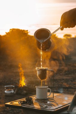 Güneşin doğuşu sırasında sarı gün ışığıyla aydınlatılan bir fincan kahve tanecikleri, kamp ateşi ve sisle çevrili el yapımı bir atmosfer ile kavrulmuş fasulye taneleri hazırlıyorum.