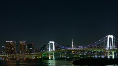 Panoramik gece manzarası, aydınlatılmış binalar, denize yansıyan uzun köprü.