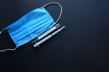 Siyah arkaplandaki tıbbi nesneler. Plastik şırınga, mavi maske ve cam cıva termometre. SARS-CoV-2 önleme.