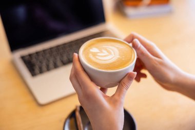 Kafede dizüstü bilgisayarın yanında elinde bir fincan cappuccino tutan genç bir kadın görüntüsü.  