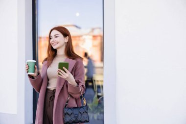 Paltolu neşeli kadın elinde kahve ile şehir sokağına gidip cep telefonunu alacak.