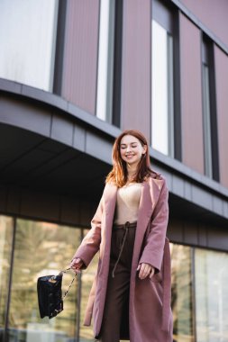 Modern binanın yanında duran el çantalı, neşeli, kızıl saçlı kadının düşük açılı görüntüsü. 