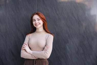 Gri mermer duvarın yanında kolları çapraz duran bej balıkçı yakalı mutlu kızıl kadın. 