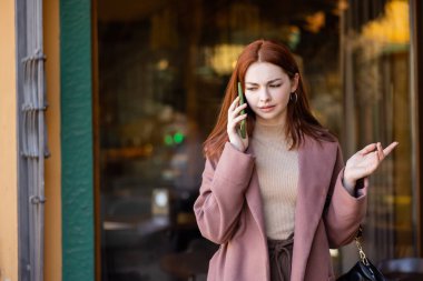 Kızıl saçlı, cep telefonuyla konuşan ve dışarıda el kol hareketi yapan genç bir kadın.