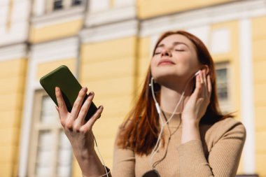 Telli kulaklıkla müzik dinleyen ve dışarıda akıllı telefon tutan memnun bir kadın. 