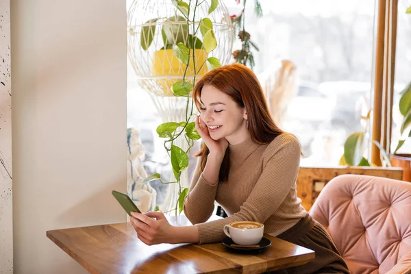 Kafede bir fincan latte yanında akıllı telefon kullanan kızıl saçlı mutlu bir kadın.