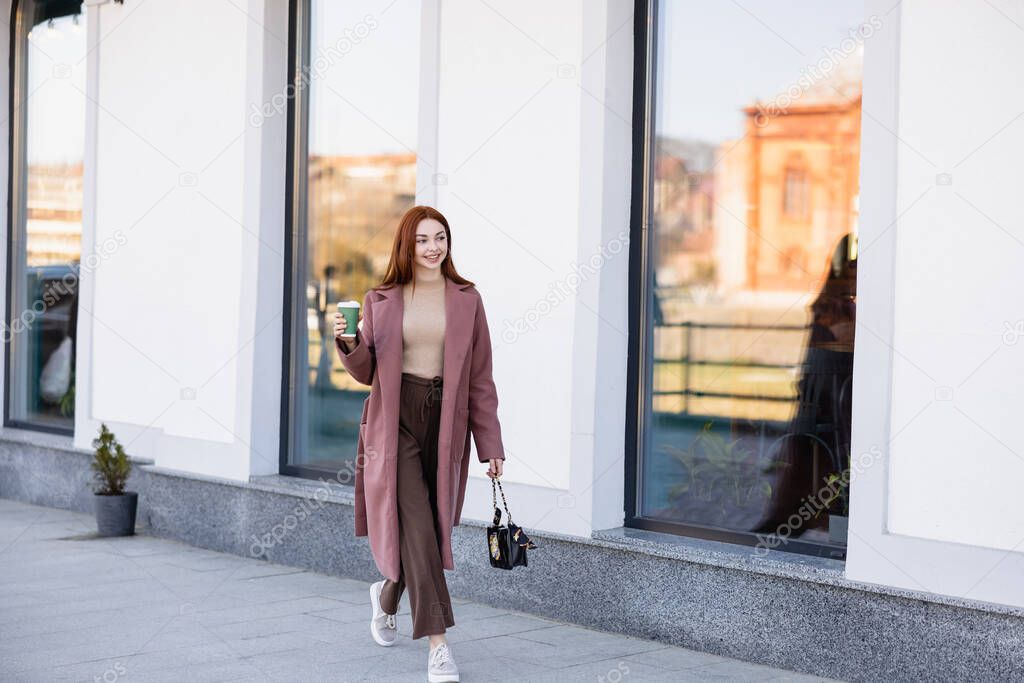 full length of pleased redhead woman with handbag holding paper cup and walking on urban street 