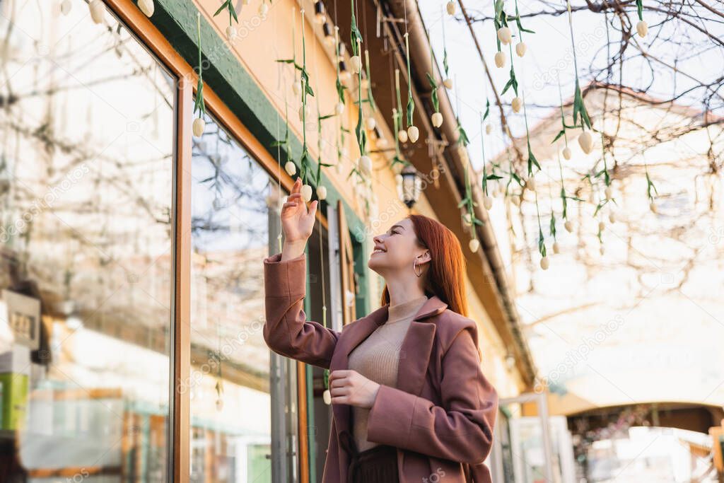 cheerful redhead woman in coat smiling while looking at hanging tulips outside 