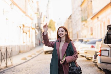 cheerful redhead woman in coat calling taxi on european street  clipart