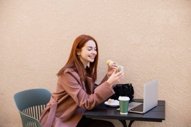 Kafe terasında dizüstü bilgisayarının yanında kızıl saçlı mutlu genç bir kadın tatlı kurabiyeler tutuyor. 