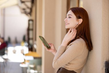 Kablosuz kulaklıklı pozitif kızıl saçlı kadın elinde akıllı telefon ve müzik dinliyor. 