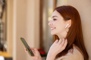 side view of positive woman in wireless earphone holding smartphone and listening music  clipart