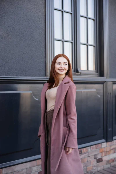身穿时髦外套 面带微笑的红头发女人站在现代建筑旁边 — 图库照片