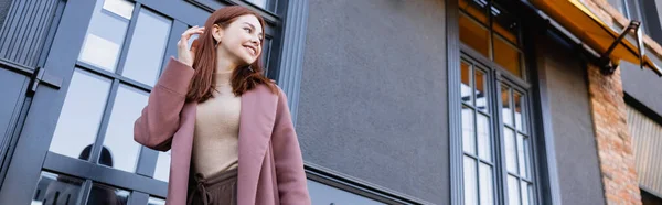 low angle view of happy woman in stylish coat adjusting red hair near modern building, banner