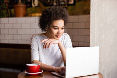 curly african american woman using laptop near cup of coffee in cafe 