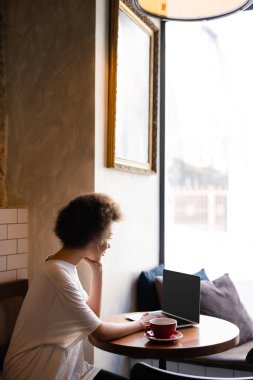 young african american woman using laptop with blank screen near cup of coffee in cafe 