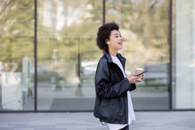 Yüzünde kulaklıkla gülümseyen Afro-Amerikan kadın. Elinde içecek ve akıllı telefon var.