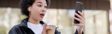 shocked african american woman in earphone holding paper cup and having video chat on smartphone, banner