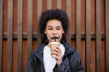 curly african american woman holding paper cup and straw with moustache near fence