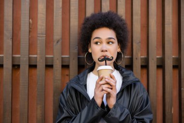 curly african american woman drinking takeaway beverage from straw with moustache near fence
