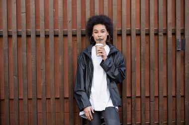 young african american woman drinking takeaway beverage from straw with moustache near fence