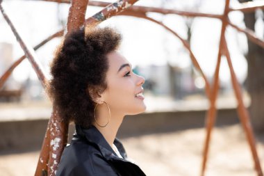 side view of happy african american woman in jacket smiling at rusty outdoor gym