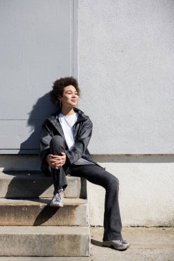 happy african american woman in jacket sitting on stairs near wall