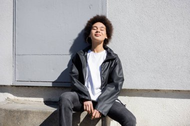 happy african american woman with closed eyes sitting on stairs near wall