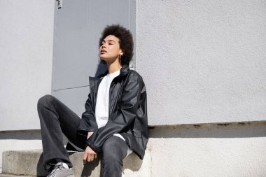 sunshine on face of stylish african american woman sitting on stairs near wall