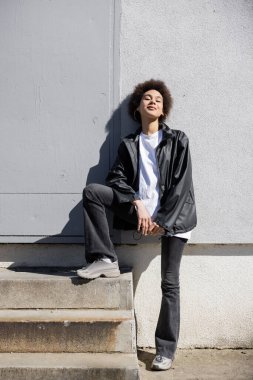 full length of stylish african american woman in jacket smiling near wall