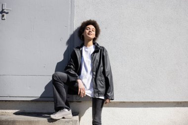 young african american woman in jacket smiling near wall