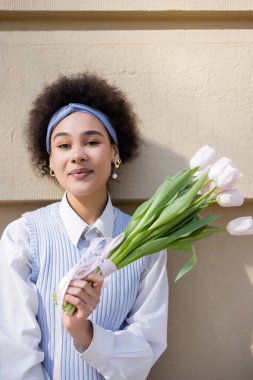 Mavi yelekli ve beyaz gömlekli mutlu Afro-Amerikan kadın. Duvarın yanında bir buket lale tutuyor.