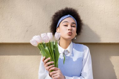 curly african american woman in blue vest and white shirt holding bouquet of tulips and pouting lips near wall clipart